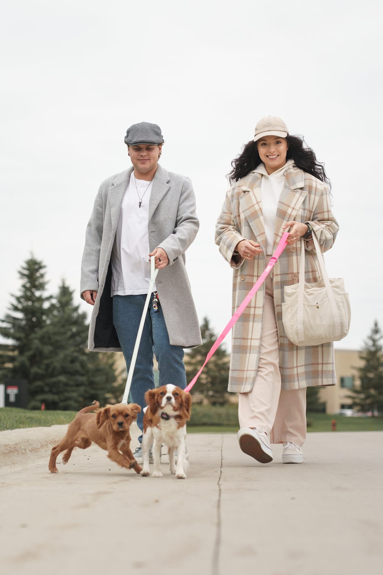 Couple in autumn coats walking two cavalier spaniels toward the camera on a park path in Winnipeg