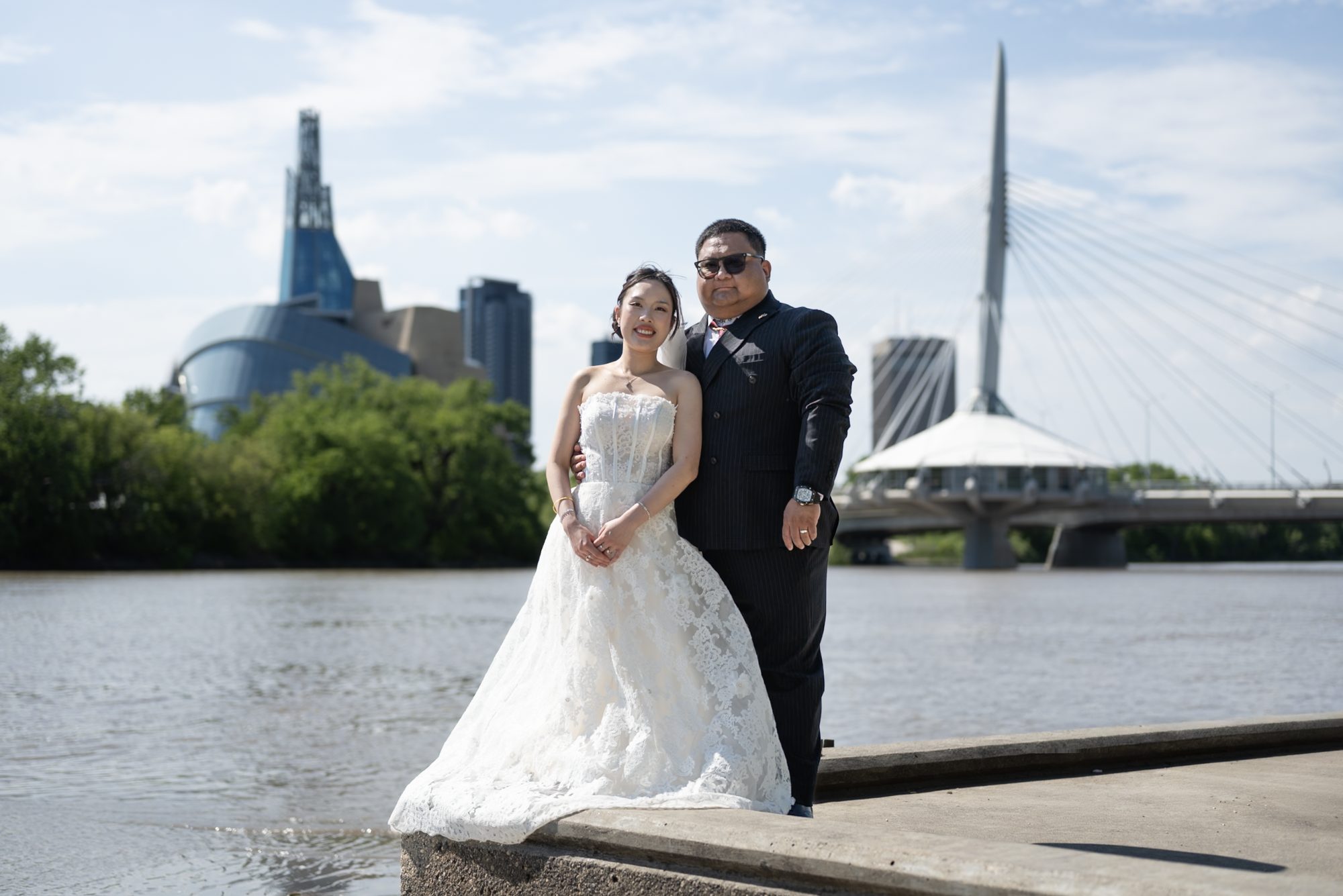 Bride and groom on the riverbank across from the St. Boniface Cathedral, with the Esplanade Riel bridge and the Canadian Museum for Human Rights behind them